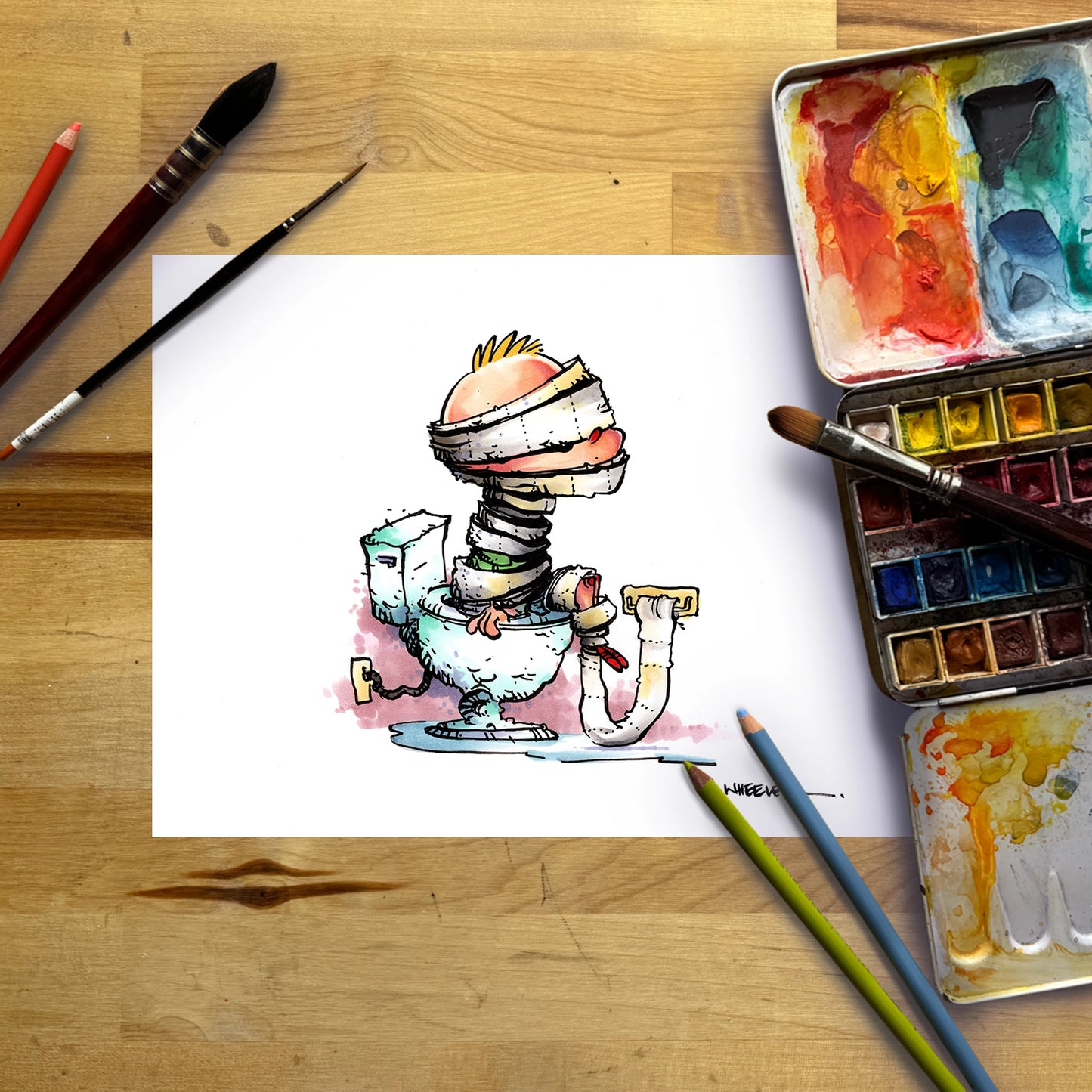 A white sheet of paper on a wooden table, featuring a whimsical cartoon drawing of a mummy-like figure sitting on a toilet. Surrounding the paper are watercolor paint palettes, paintbrushes, and colored pencils.