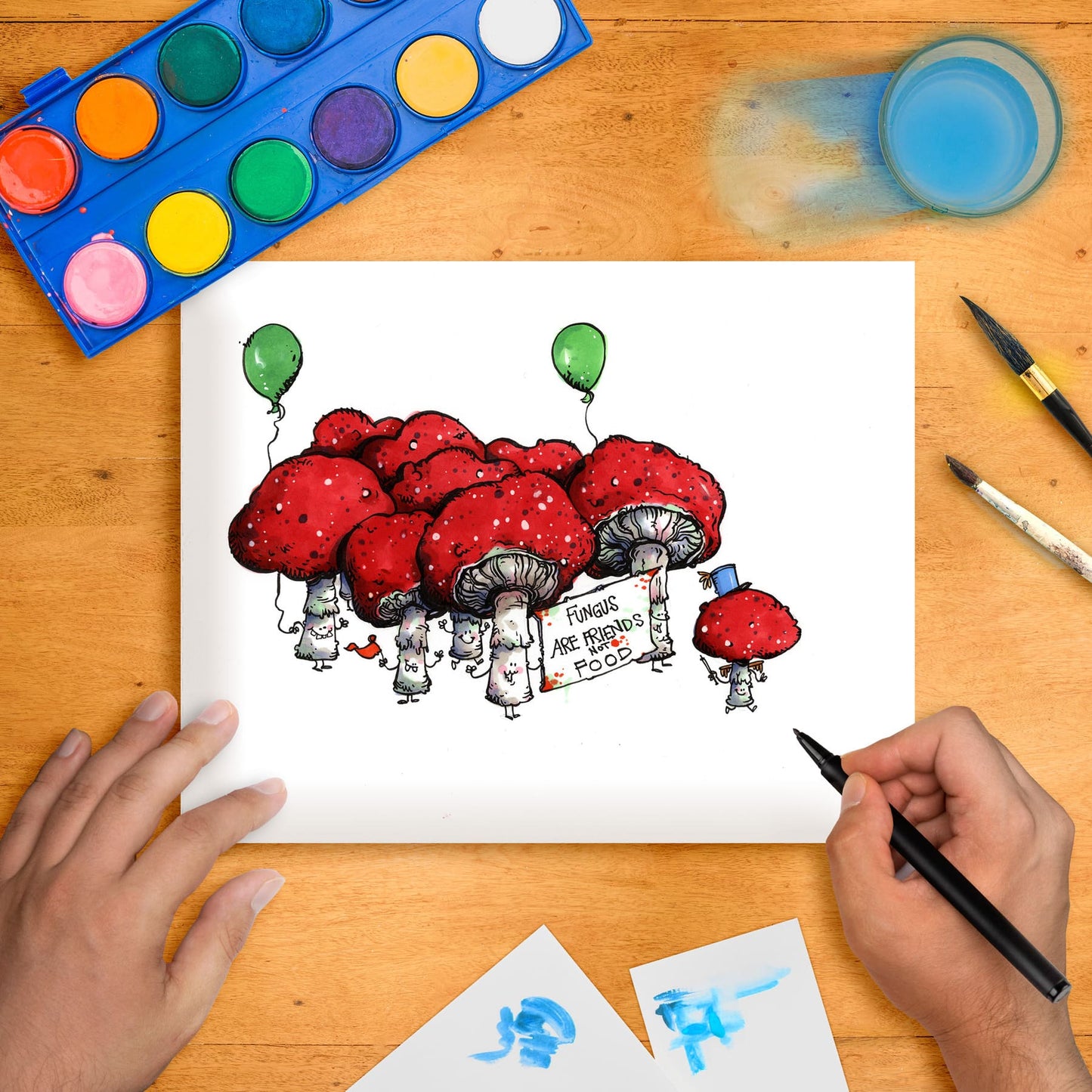 Original marker illustration artwork on paper depicting whimsical red mushrooms with eyes and hats, titled 'Fungus Parade'. The artwork is shown with a person holding a marker over it, surrounded by art supplies like paint and brushes on a wooden table.