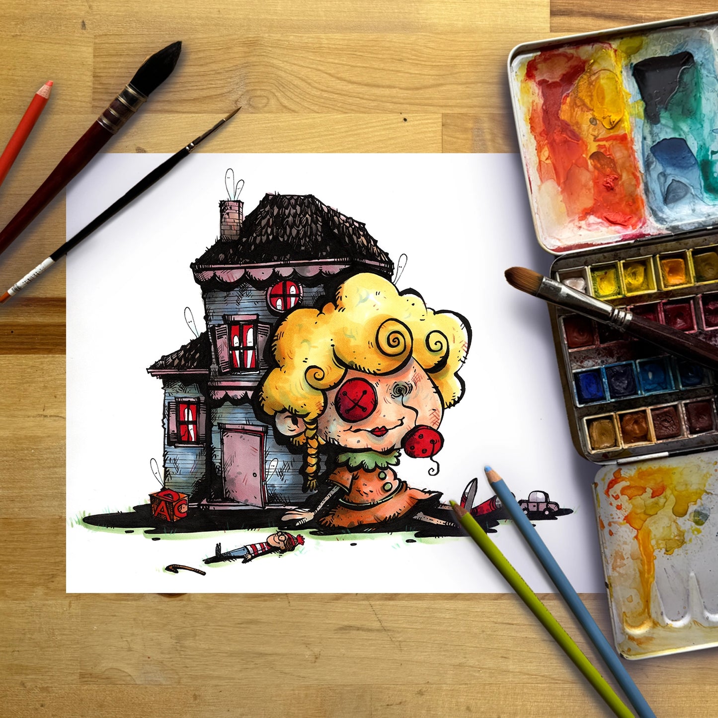 A whimsical hand-drawn illustration of a clown-like character with curly yellow hair and red eyes sitting in front of a dark haunted house, shown on a white sheet of paper surrounded by watercolor paints, brushes, and pencils on a wooden table.