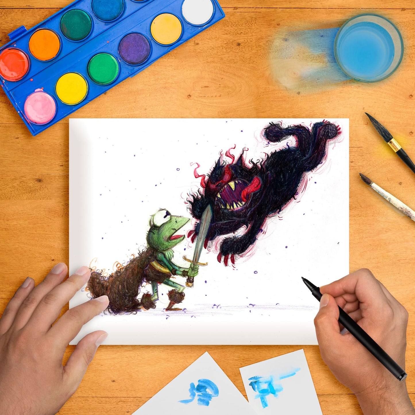 Original marker illustration artwork on paper depicting a cartoon-style frog character interacting with a fantastical, black, cat-like creature. The artwork is accompanied by various art supplies like paint, brushes, and markers on a wooden table.