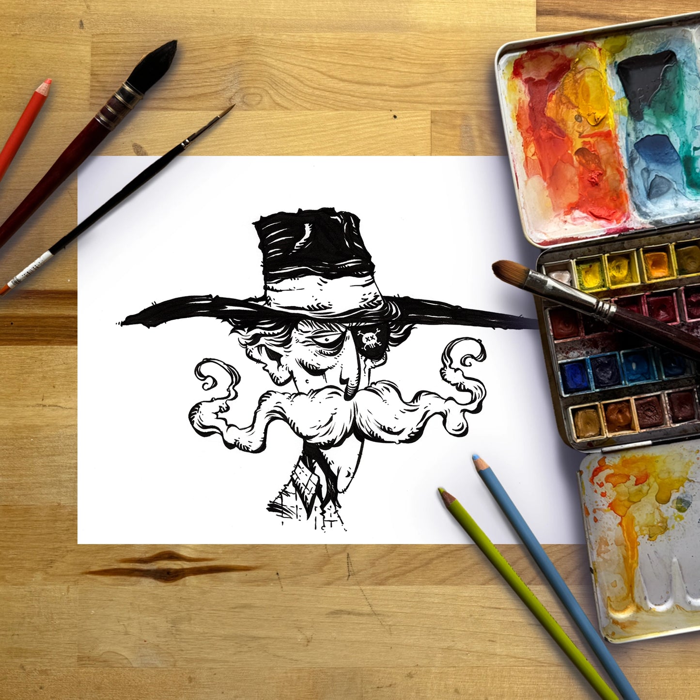 A black ink sketch of a stylized face with a large hat and curled mustache on a white sheet of paper, arranged on a wooden tabletop with paintbrushes, colored pencils, and watercolor paint palettes.