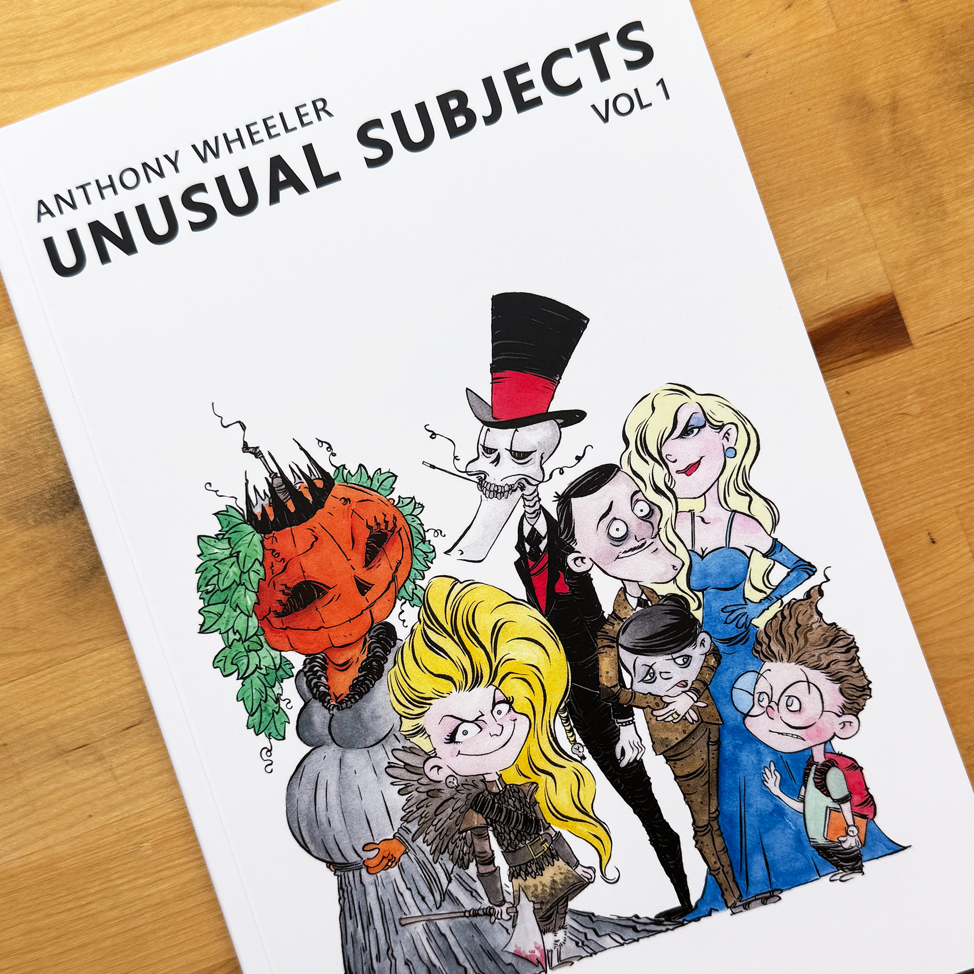 Anthony Wheeler’s Unusual Subjects Vol. 1 book cover featuring a colorful group of unusual illustrated characters on a wooden tabletop.