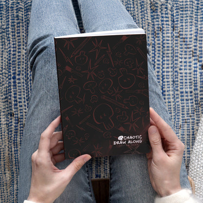 Person holding the black Chaotic Draw Along Sketchbook while seated on a blue patterned rug.