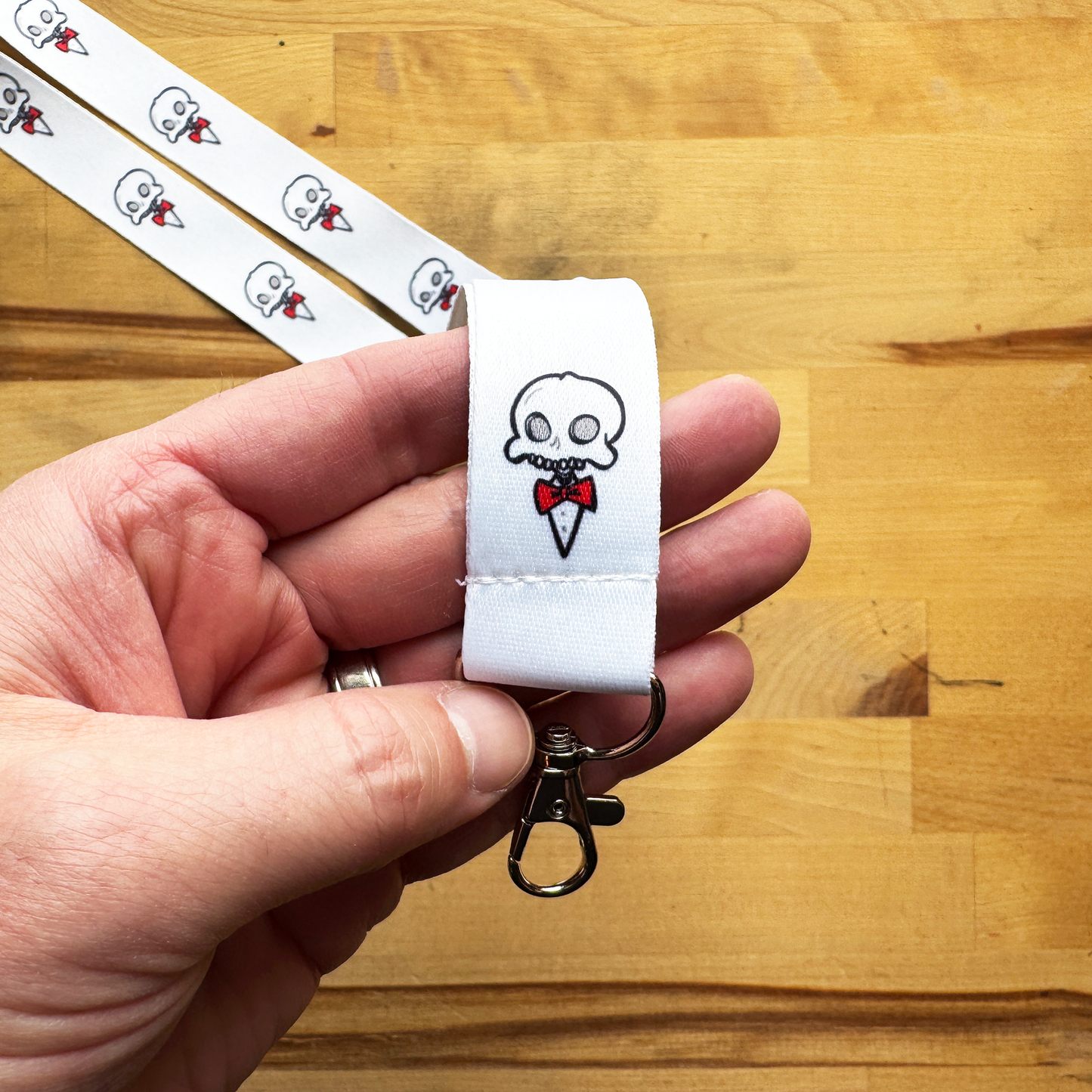 White Chaotic Draw Along lanyard keychain with skull and red bow tie design, held above a wooden tabletop.