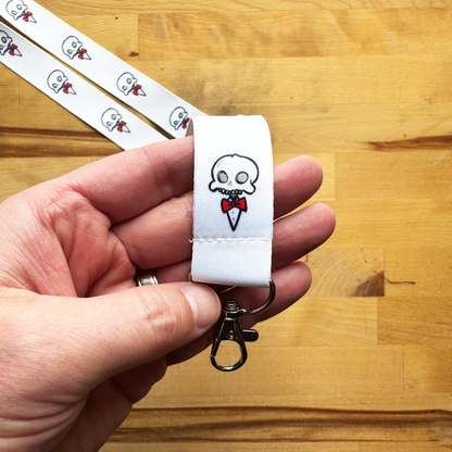 White Chaotic Draw Along lanyard keychain with skull and red bow tie design, held above a wooden tabletop.