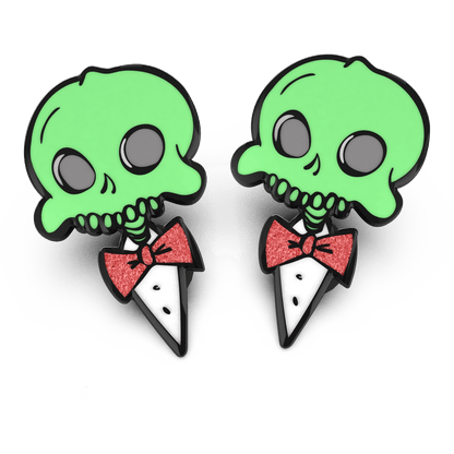 Two enamel pins with a glow in the dark effect, featuring a character with a skeleton grin and a red bow tie.