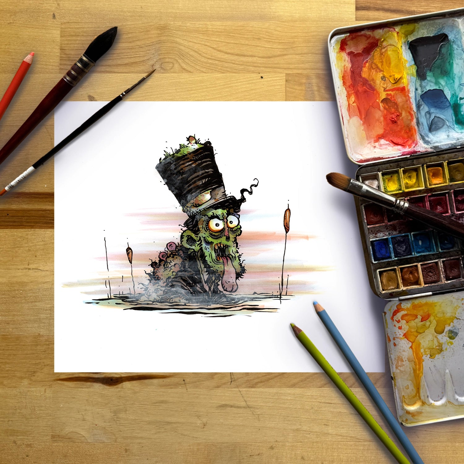 A watercolor painting of a green zombie-like character wearing a tall top hat, shown on a sheet of white paper surrounded by art supplies including paintbrushes, watercolor paints, and colored pencils on a wooden surface.