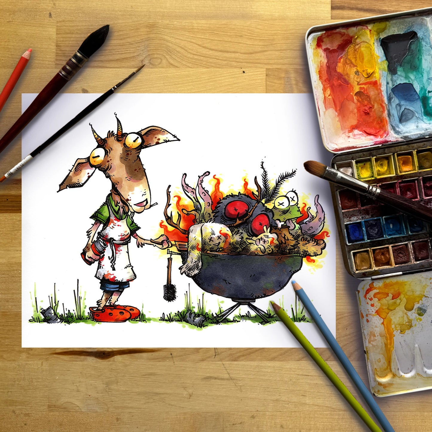 A cartoon-style watercolor illustration on a white sheet of paper, showing a goat-like character standing beside a cauldron filled with colorful monsters and flames. The paper is photographed on a wooden surface with paintbrushes, colored pencils, and watercolor palettes around it.