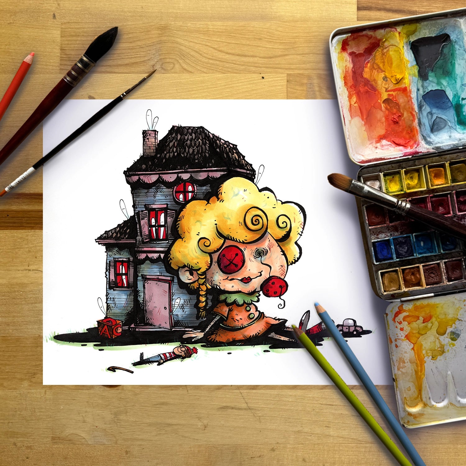 A whimsical hand-drawn illustration of a clown-like character with curly yellow hair and red eyes sitting in front of a dark haunted house, shown on a white sheet of paper surrounded by watercolor paints, brushes, and pencils on a wooden table.