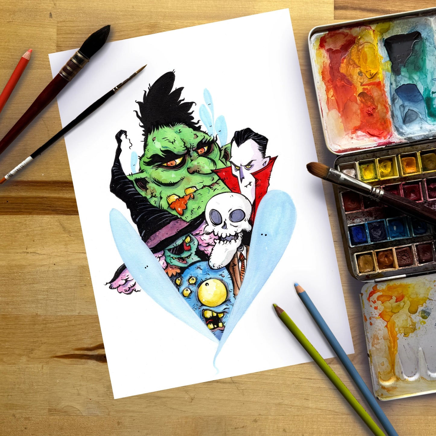 A colorful watercolor illustration of a green monster and a vampire with a skull, shown on a white sheet of paper surrounded by paintbrushes, watercolor paints, and colored pencils on a wooden surface.