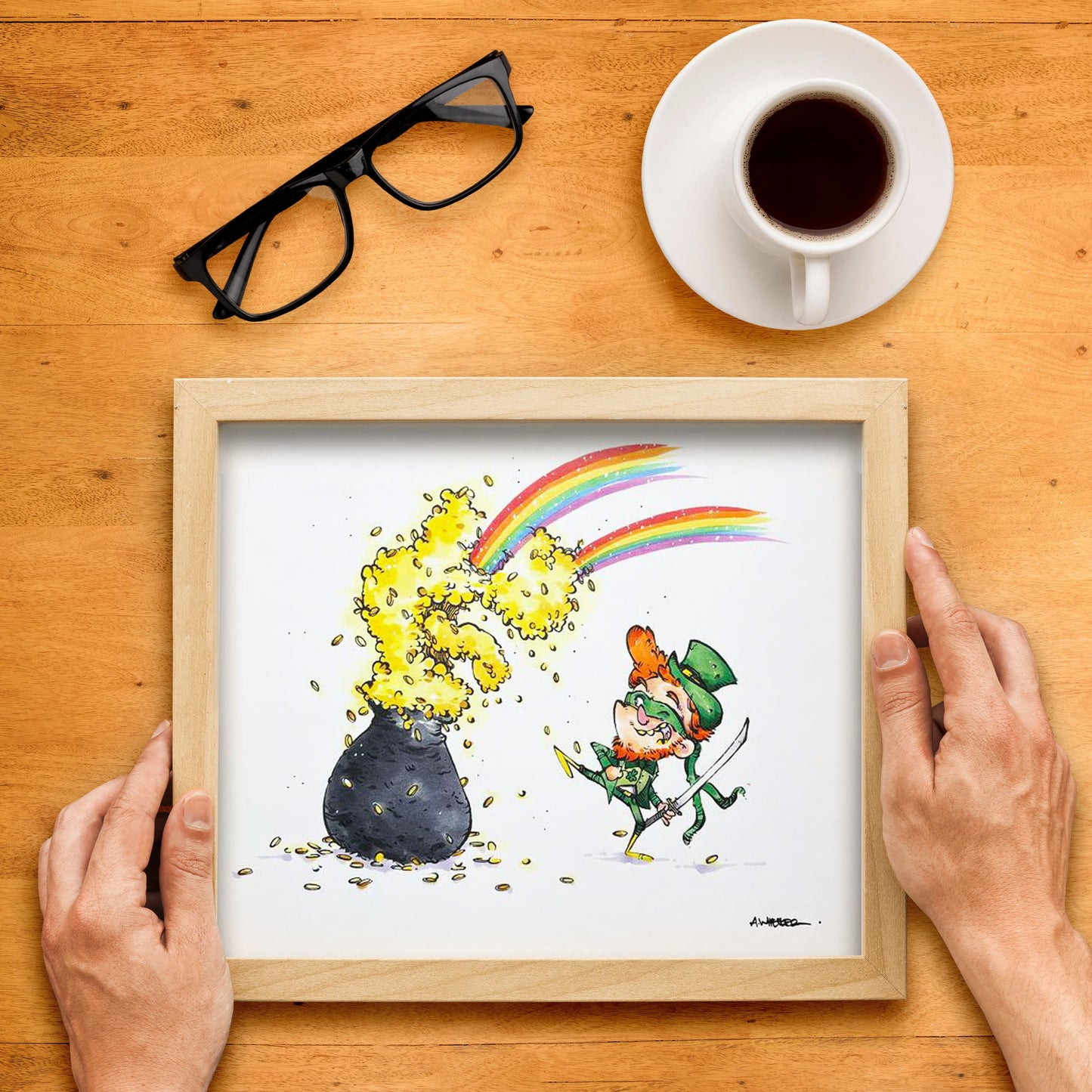 Original artwork depicting a cartoon-style leprechaun with a pot of gold, with a rainbow-colored backdrop, held in front of a wooden frame and a cup of coffee.