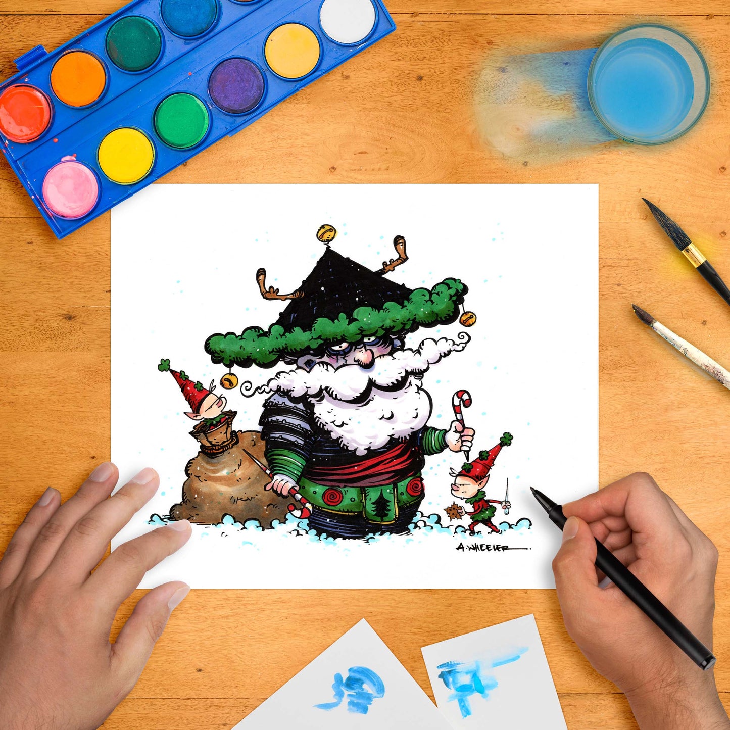 Festive artwork of a stylized Santa dressed as a samurai warrior, flanked by mischievous elves, with drawing tools and a paint palette arranged around the hand-colored print on a wooden desk.