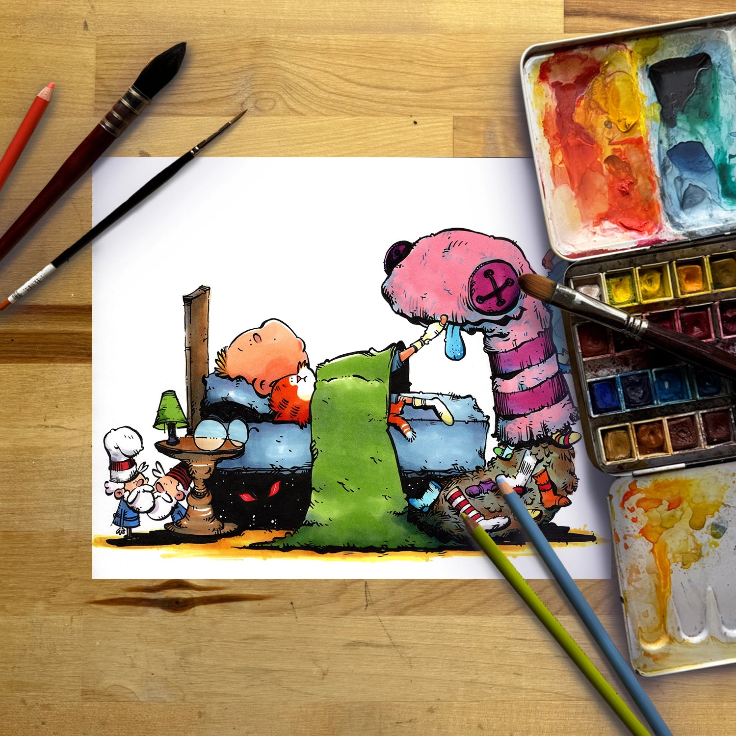 A colorful cartoon illustration printed on a white sheet of paper, laid flat on a wooden tabletop with paintbrushes, a watercolor palette, and pencils around it.