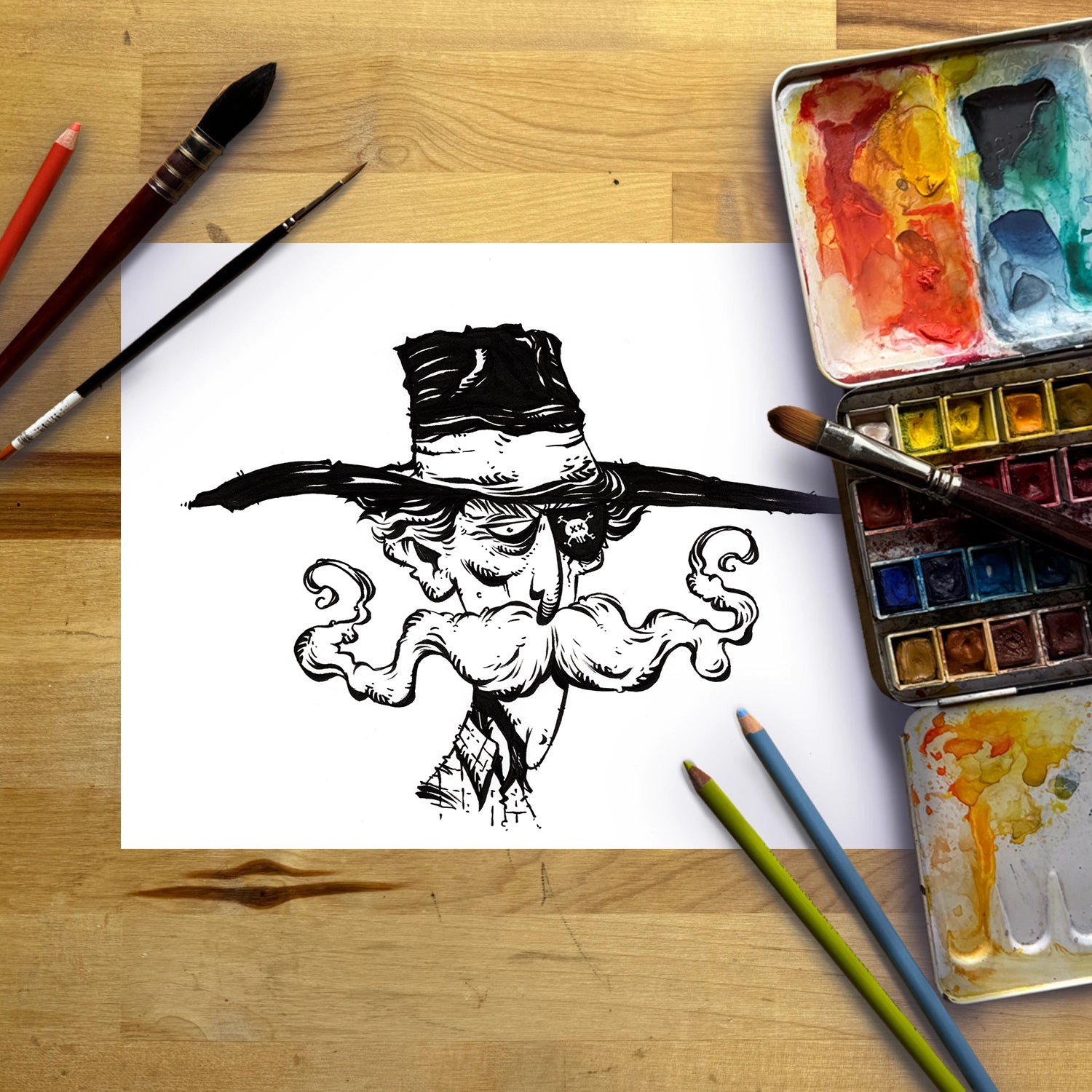A black ink sketch of a stylized face with a large hat and curled mustache on a white sheet of paper, arranged on a wooden tabletop with paintbrushes, colored pencils, and watercolor paint palettes.