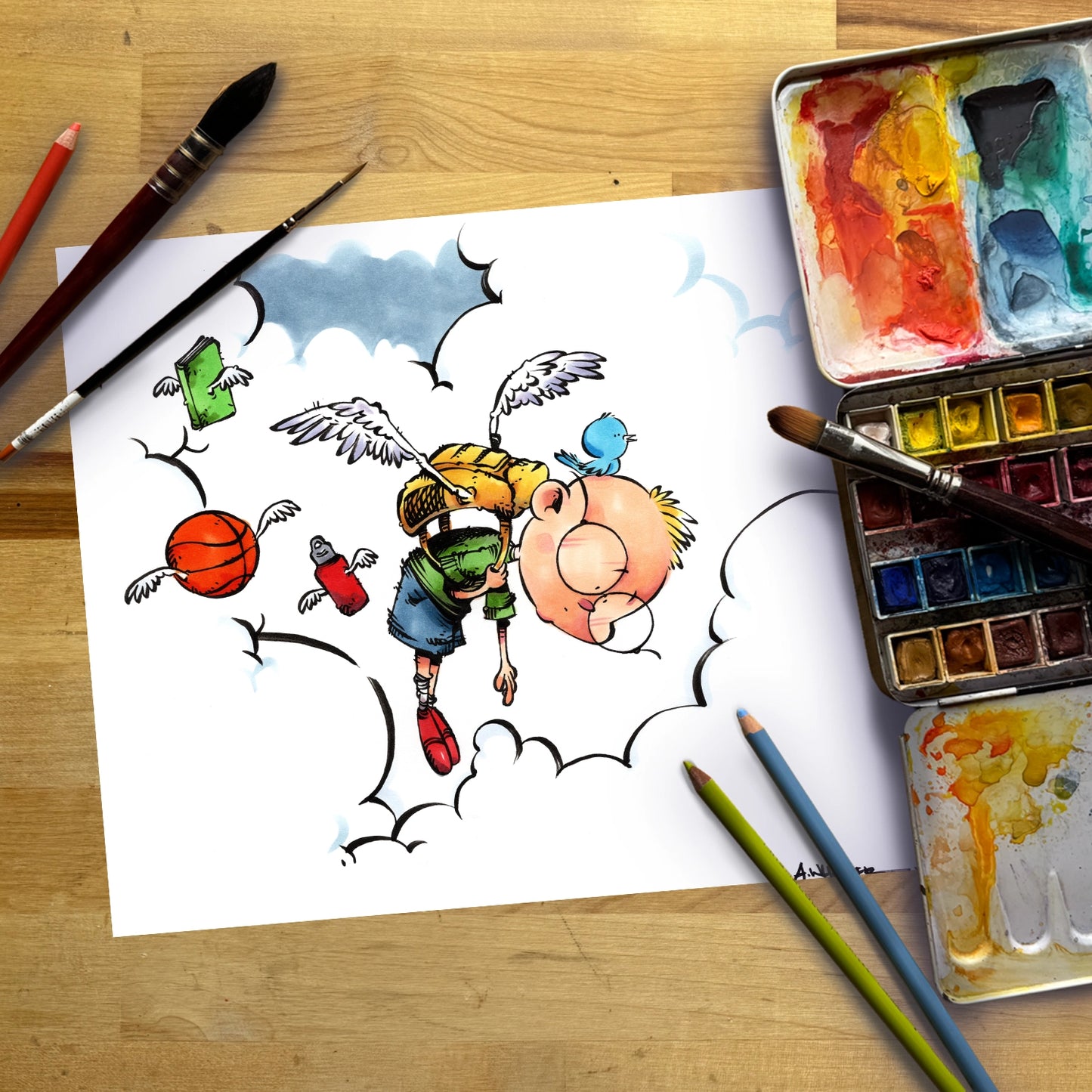 A flat-lay photograph of a white sketchbook on a wooden table, featuring a cartoon illustration of a winged character with a basketball and a blue bird. Surrounding the sketchbook are watercolor paint palettes, paintbrushes, and colored pencils.