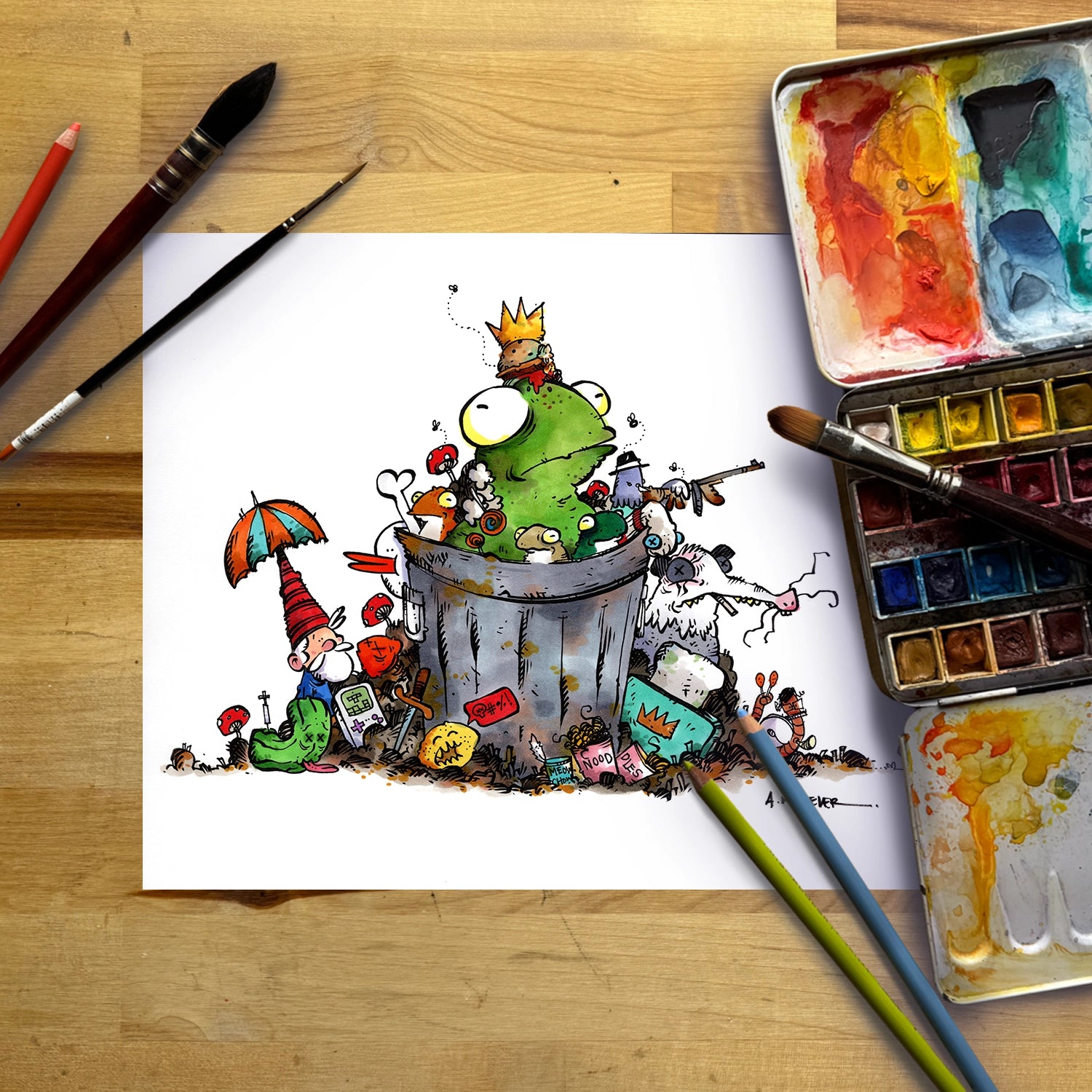 A whimsical cartoon illustration on a sheet of white paper, showing a green frog-like character sitting in a trash can with a small gnome and colorful trash elements. The paper is photographed from above on a wooden table with paintbrushes, a watercolor palette, and two paint sticks around it.